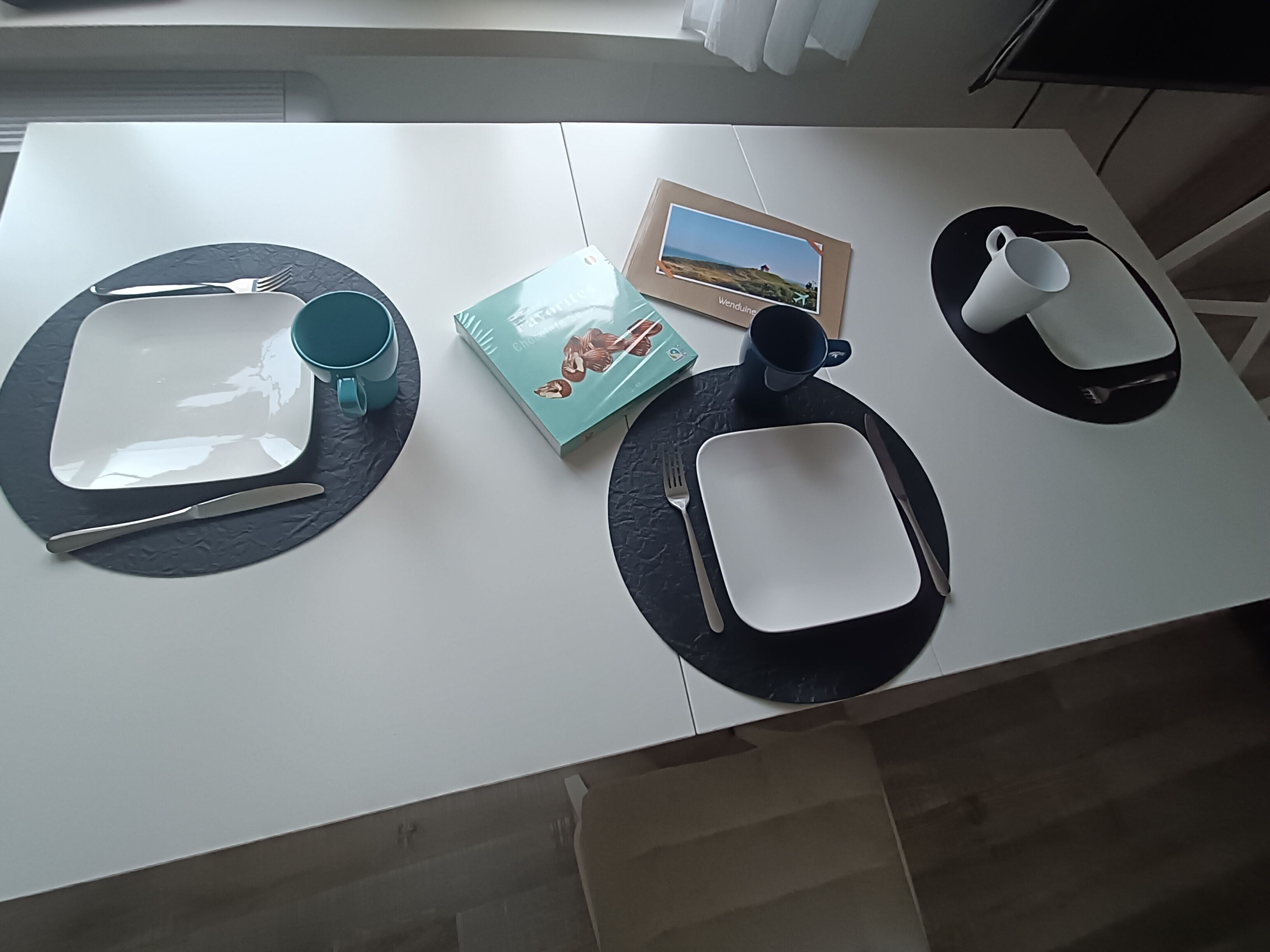Ein Esstisch mit weißer Tischdecke, schwarzen Tischsets, einem Buch, einem gerahmten Foto, einer Tasse und einem Teekännchen.