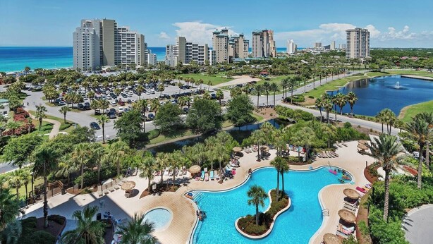 Un resort con varios edificios de gran altura, un amplio estacionamiento y una piscina central rodeada de palmeras y zonas de descanso.