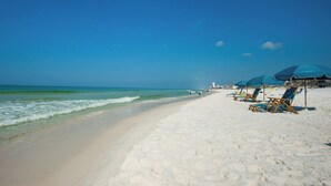 Una playa de arena blanca y agua azul cristalina. Se observan sombrillas azules y sillones de madera a lo largo de la orilla.