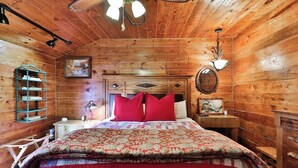 Habitació d'una cabana de fusta amb un llit, una tauleta de nit, una làmpada, una petita estant i una prestatgeria penjada a la paret.
