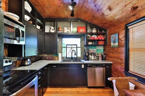 A kitchen with wooden cabinets, a stainless steel dishwasher, and a variety of dishes and kitchenware on shelves. - The Hayloft - A Sayles Ranch Home (Abilene)