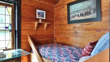 A cozy cabin room with a wooden ceiling, a bed with plaid bedding, a small table with a bowl, a framed picture on the wall, and a window with a stained glass design.