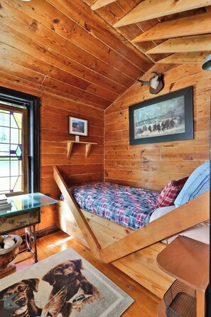 Ein gemütliches Blockhauszimmer mit Holzbalkendecke, einem Bett mit Karo-Bettwäsche, einem kleinen Tisch mit einer Schale, einem gerahmten Bild an der Wand und einem Fenster mit Buntglasdekor.