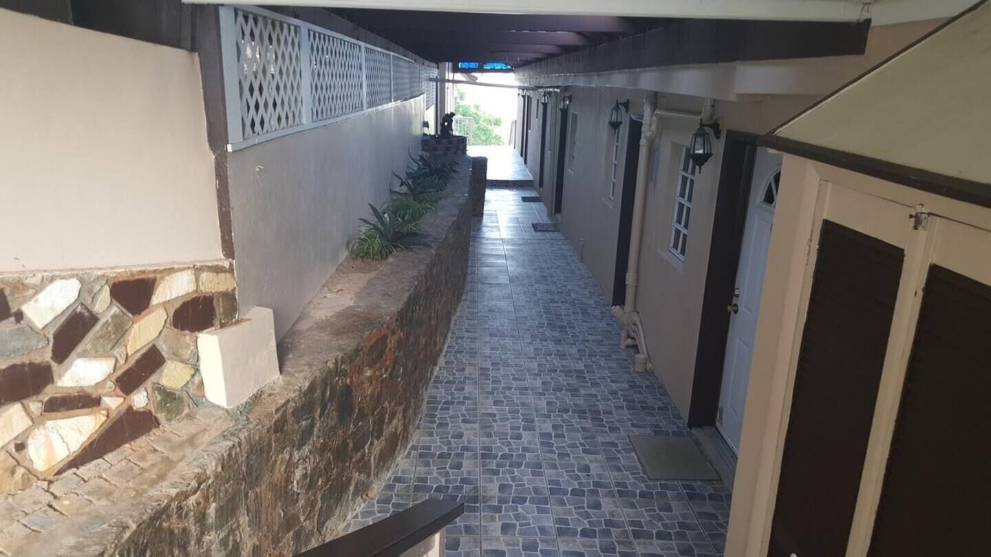 A narrow corridor with a stone wall on one side and a tiled floor. There are doors on the right and a gate at the end.