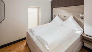 Ein ordentlich bezogenes Bett in einem Hotelzimmer mit einem Holzkopfteil, einem kleinen Nachttisch mit Fernbedienung und einem Fenster mit Jalousien.