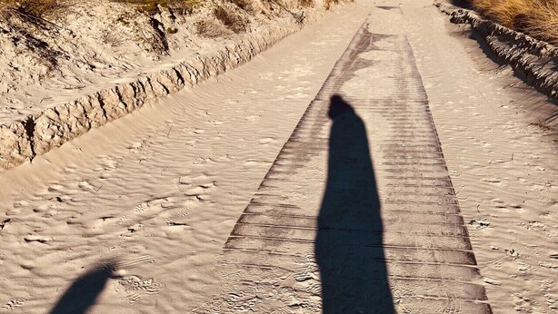 Eine lange, gerade Straße, begleitet von grasbewachsenen Dünen, mit dem Schatten einer wandernden Person.