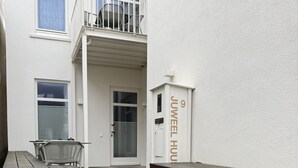Ein moderner Außenbereich mit einer Holzbank, einem Metallstuhl und einem kleinen Tisch. Eine Treppe aus Holz führt zu einer Tür mit Glasfenster.