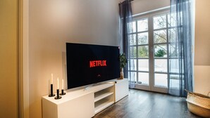 Ein modernes Wohnzimmer mit einem großen Flachbildfernseher auf einem weißen Standfuß, einem Fenster mit transparenten Vorhängen und einem Holzboden.