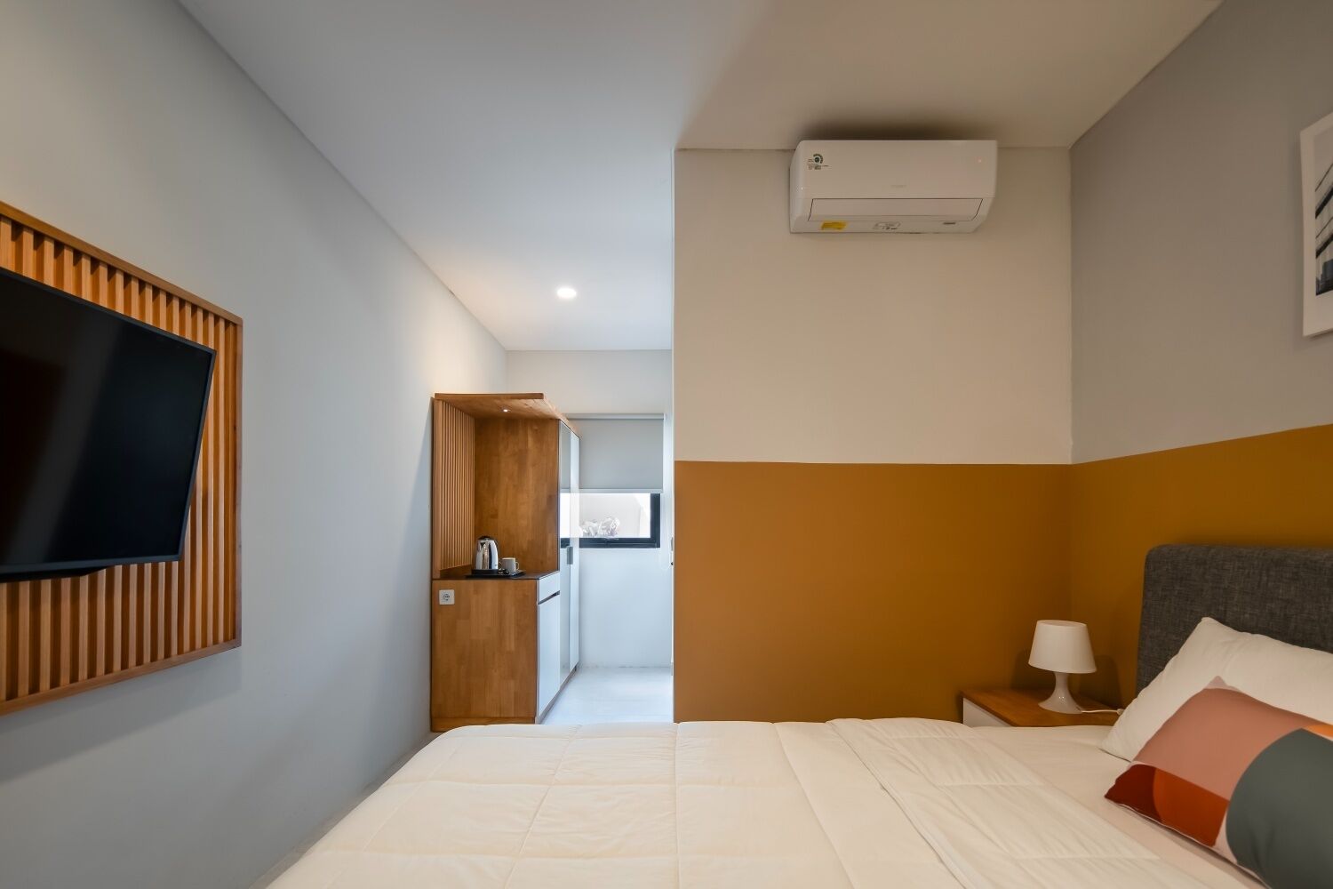 A hotel room with a bed, a TV mounted on the wall, a small bedside table with a lamp, and a window.