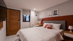 A bedroom with a bed, a wooden wardrobe, a bedside table with a lamp, and a small shelf with decorative items. - Cove Urbanier (Jakarta)