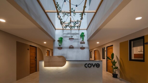 A reception desk with a wooden top and a 'cove' sign, flanked by wooden doors and potted plants. - Cove Urbanier (Jakarta)