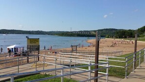 Une plage avec un panneau indiquant la présence d’un sauveteur, un panneau de signalisation pour le stationnement et une zone délimitée par une clôture et des garde-corps.
