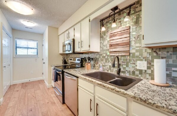A kitchen with white cabinets, a granite countertop, a double sink, a microwave, and a tiled backsplash with a phrase.