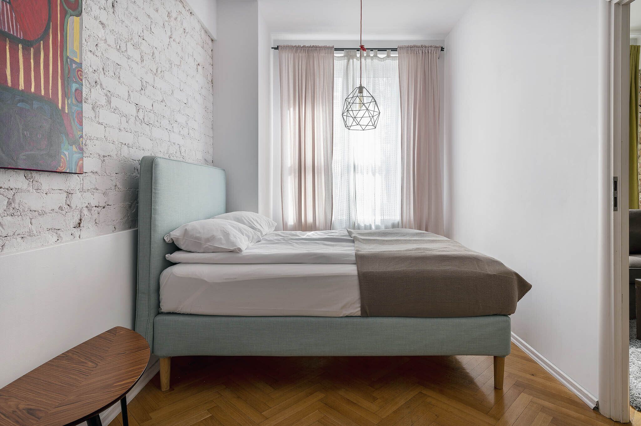 Un dormitorio con cama, una mesita, lámpara colgante y ventana con cortinas.