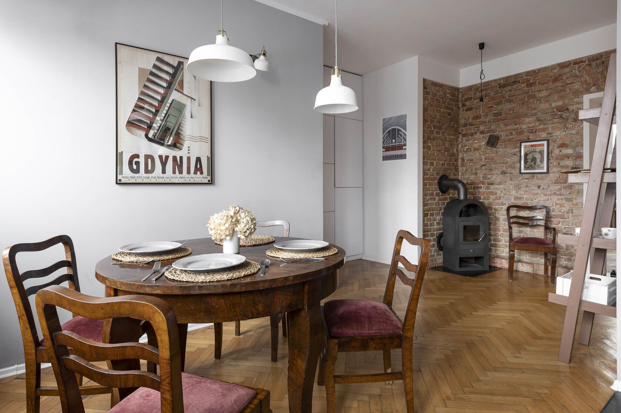 A dining area with a wooden table, chairs, and pendant lights. There is a brick wall and a fireplace in the background.