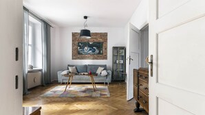 A living room with a herringbone wooden floor, a grey sofa with cushions, a coffee table, a hanging light fixture, and a brick wall with a painting. - Roxy by Q4Apartments (Gdynia)