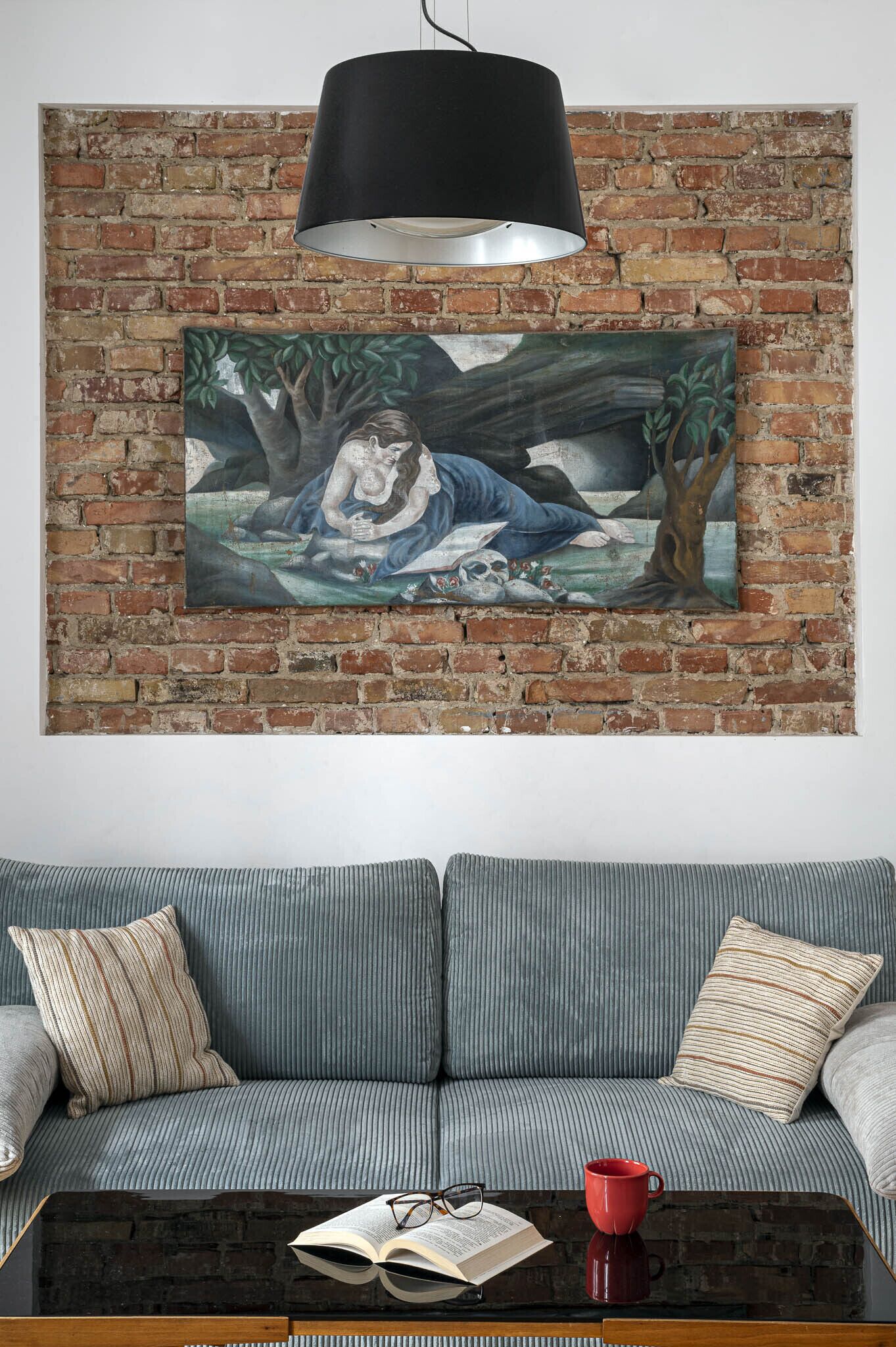 A living room with a brick wall, a large painting, a grey sofa, and a coffee table with a book and a red mug.