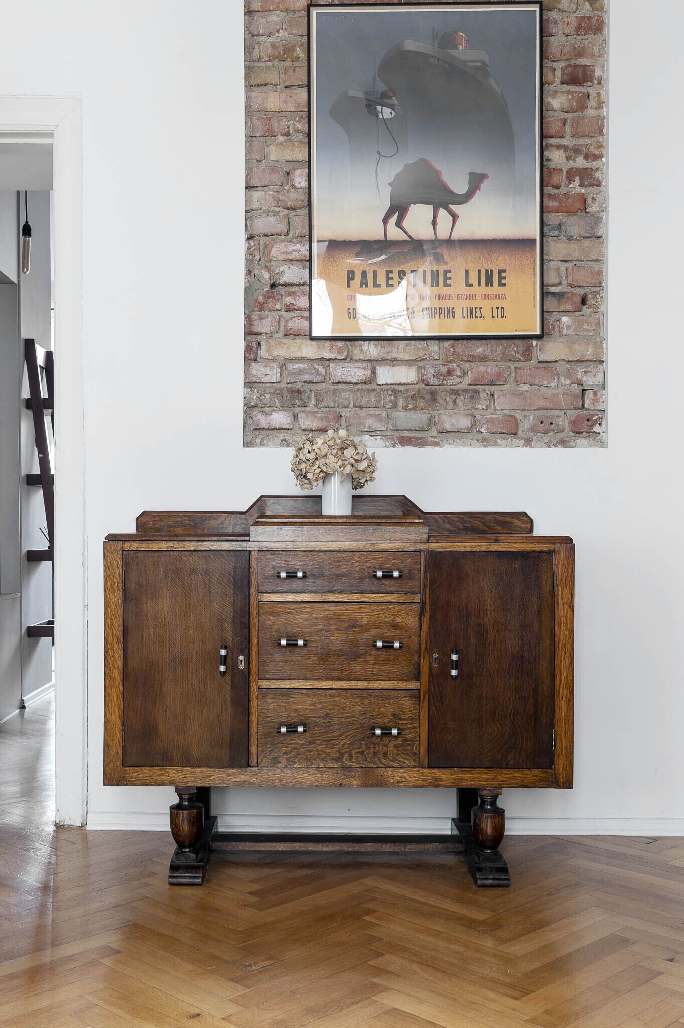 A wooden cabinet with drawers and a vase with dried flowers on top, placed against a brick wall with a poster above it.