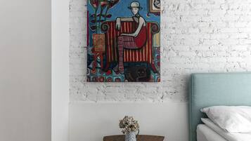Una habitación con pared de ladrillo blanco, una cama con cabecera color turquesa, una pequeña mesa de madera con un jarrón de flores secas y un cuadro abstracto y colorido sobre la mesa.