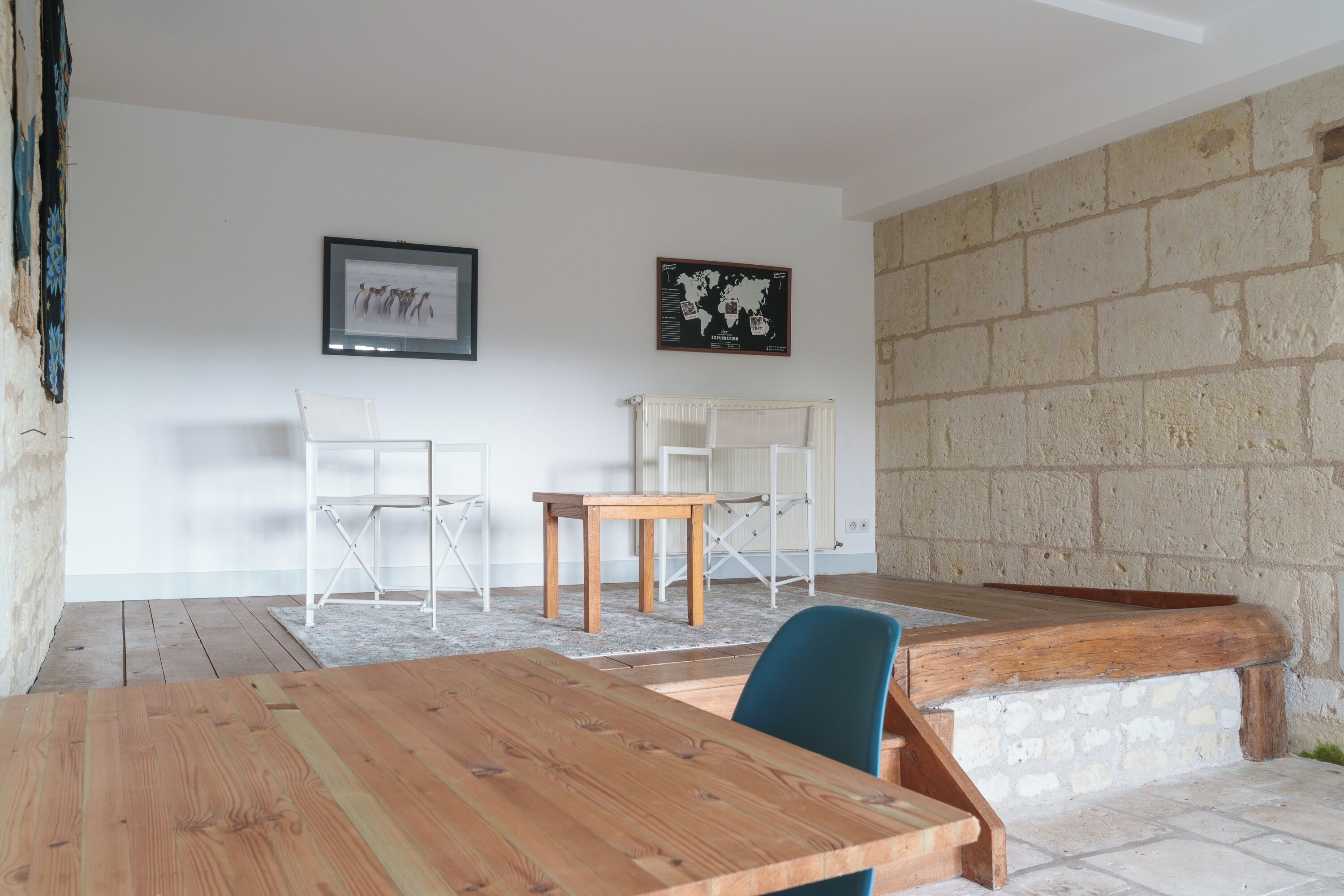 Une pièce avec une table en bois, deux chaises blanches, une petite table en bois et un mur en pierre.