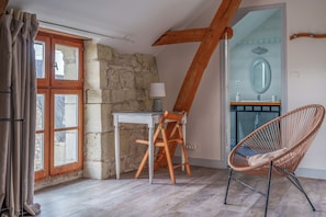 Ein Zimmer mit Steinwand, Holzbalken, einem Tisch mit Stuhl, einem Korbstuhl und einem Badezimmer mit Spiegel und Waschbecken.