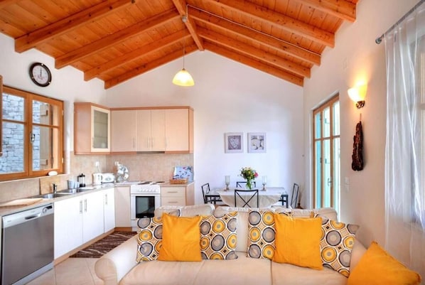 Un intérieur chaleureux avec un plafond en bois, une cuisine dotée de meubles blancs, un coin repas avec une table et des chaises, et un canapé agrémenté de coussins à motifs.