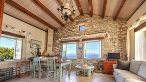 A rustic interior with a stone wall, wooden beams, a dining area with chairs and a table, a sofa, and a television. - Villa Agathi - Two Bedroom Villa, Sleeps 4 (Paxi)