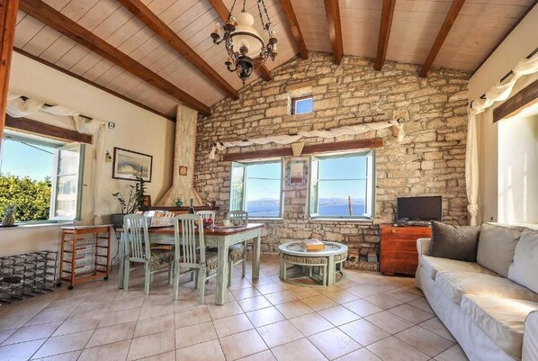 A rustic interior with a stone wall, wooden beams, a dining area with chairs and a table, a sofa, and a television. - Villa Agathi - Two Bedroom Villa, Sleeps 4 (Paxi)