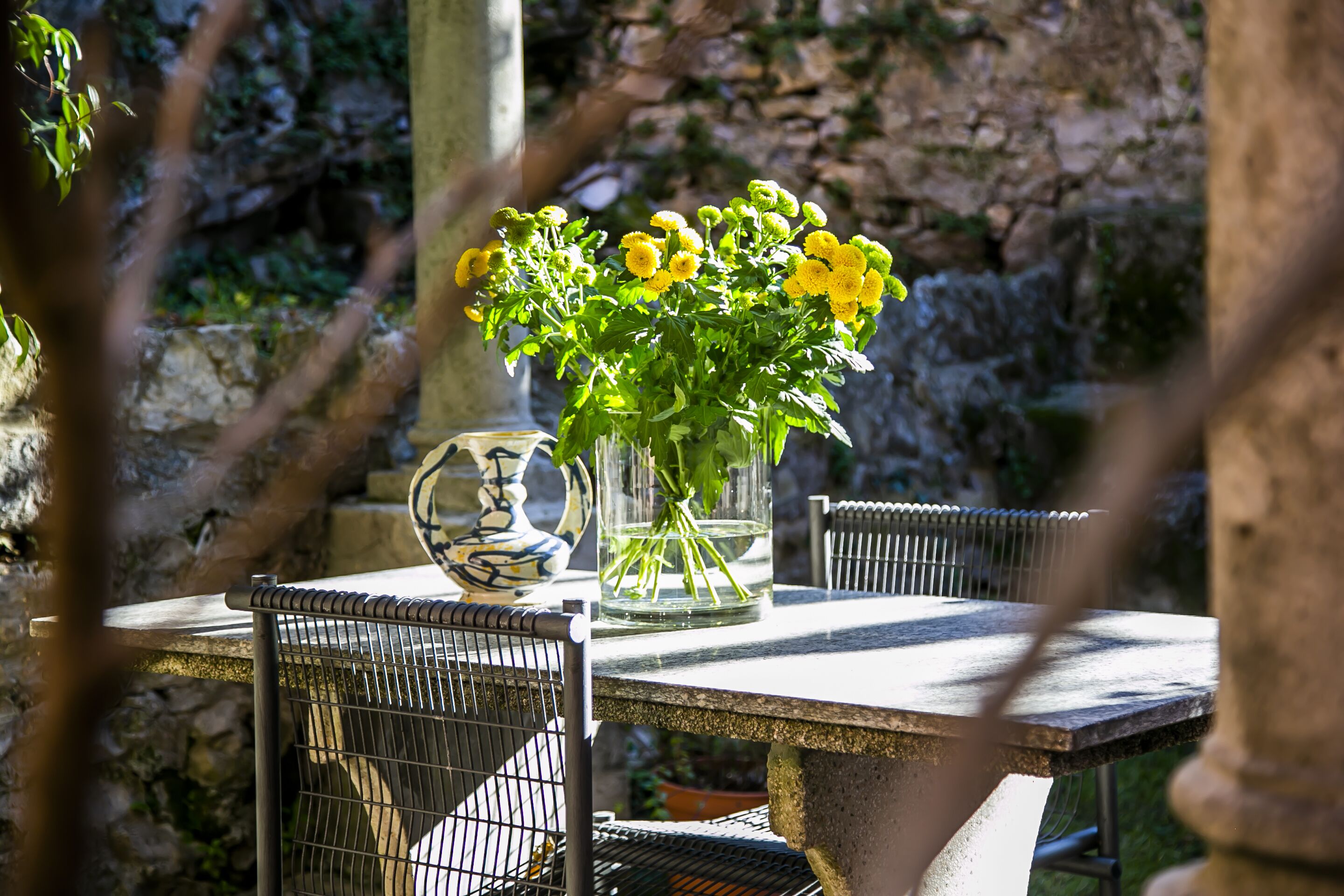 A table with a vase of yellow flowers, a decorative bowl, and metal chairs.