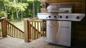 A stainless steel grill with four burners, a side burner, and a storage cabinet, set on a wooden deck with a wooden railing. - Spacious Family Cabin, Hot Tub & Pet Friendly! (Cobourg)