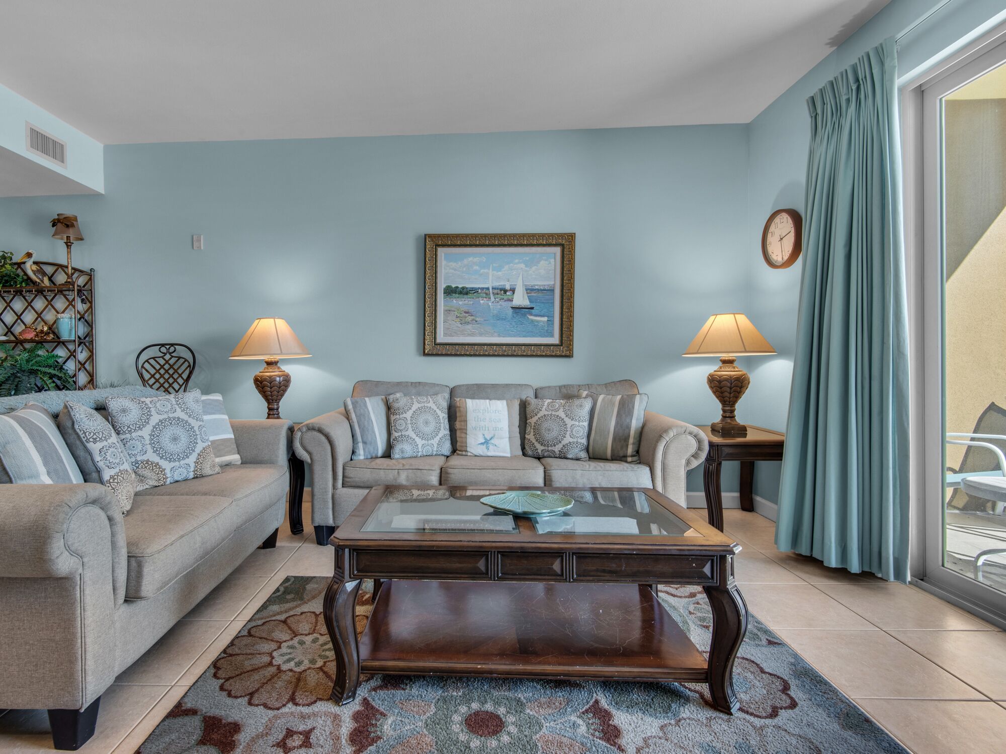 A living room with two sofas, a coffee table, and two lamps. There is a painting on the wall and a clock on the wall.