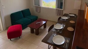 A compact living space with a sofa, a red ottoman, a dining table set for four, and a TV mounted on the wall. - Gîte Morne aux Vents (Case-Pilote)