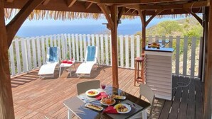 A covered outdoor dining area with a view of the ocean, wooden deck, white fence, and lounge chairs. - Gîte Morne aux Vents (Case-Pilote)