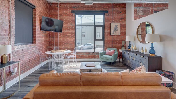 A modern living room with a brick wall, a large window, a sofa, a dining area, and a TV mounted on the wall. - The Hive 103 Bees Knees Pet Friendly by BlueStar (Wilmington)