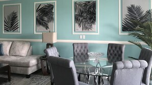 Un salon avec un mur de couleur turquoise, une table basse en verre, des fauteuils à revêtement gris et des œuvres d’art représentant des feuilles, encadrées.