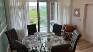Un salon spacieux avec une table en verre entourée de chaises et d’un canapé, et une vue sur un paysage verdoyant grâce à de grandes fenêtres.