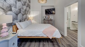 A bedroom with a bed, a nightstand, a wall-mounted lamp, a framed picture, and a mirror. - The Hive 105 Bees Knees by BlueStar (Wilmington)