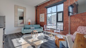 A modern living room with a brick wall, a large window, a sofa, a coffee table, and a TV. - The Hive 105 Bees Knees by BlueStar (Wilmington)