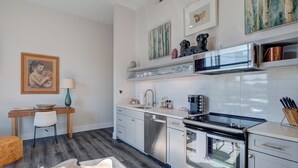 A modern kitchen with white cabinets, a stainless steel dishwasher, a built-in oven, and a microwave. There is a wooden desk with a chair and a lamp on the left side. - The Hive 105 Bees Knees by BlueStar (Wilmington)