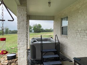 A hot tub with steps, a window, and a hanging light fixture in a covered outdoor area with a brick wall. - Casa Blanca: Hot-Tub & Firepit w/Hill Country View (Fredericksburg)