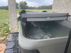 A hot tub with a cover, set against a brick wall, outdoors with a grassy field in the background. - Casa Blanca: Hot-Tub & Firepit w/Hill Country View (Fredericksburg)