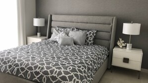 Un dormitorio moderno con un cabecero tapizado gris, una colcha estampada, dos mesitas de noche con lámparas y un pequeño objeto decorativo sobre una de ellas.