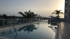 Una piscina en la azotea con paredes de vidrio transparente, palmeras y el perfil de una ciudad al fondo.