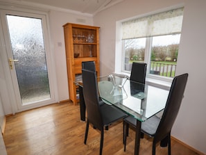 A dining area with a glass table, black chairs, a wooden cabinet with glassware, and a view through a window. - Wayside Lodge (Craven Arms)