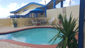 Une piscine avec, en arrière-plan, un bâtiment aux couleurs bleu et blanc, une table avec des chaises et des plantes en pot.