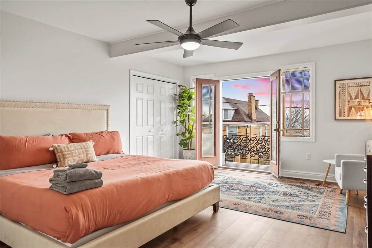 Een slaapkamer met een bed, een plafondventilator, een deur naar een balkon en uitzicht op een huis.