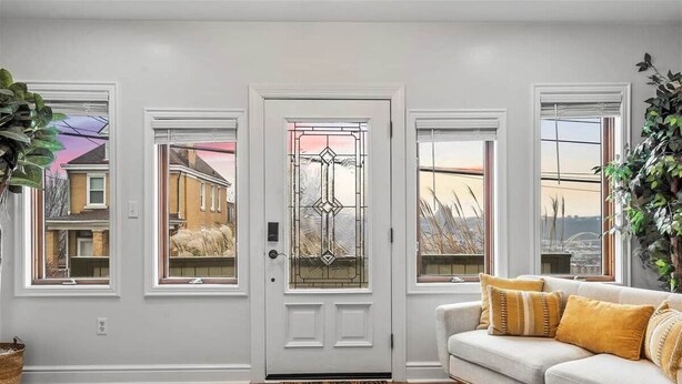 A room with a white door, a beige sofa with yellow cushions, and a view of a residential area through the windows. - Fine View | Rooftop Deck | 4BR (Pittsburgh)