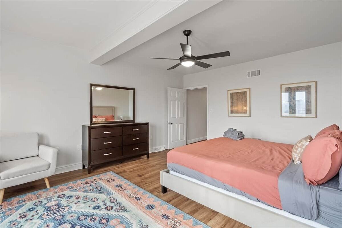 Une chambre à coucher comprenant un lit, une commode, une chaise, un ventilateur de plafond et un tapis à motifs.