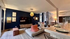 Una sala moderna con una pared de acento azul oscuro, un televisor de pantalla plana grande, un mueble de entretenimiento de madera, una escalera de madera redonda y una zona de comedor con una mesa y sillas de madera.