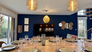 Un comedor con una mesa de madera dispuesta para seis personas, dos lámparas colgantes, un televisor de pantalla plana grande y una pared de acento azul oscuro.